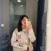 A woman takes a mirror selfie in a bathroom, wearing a beige jacket with black and orange details.