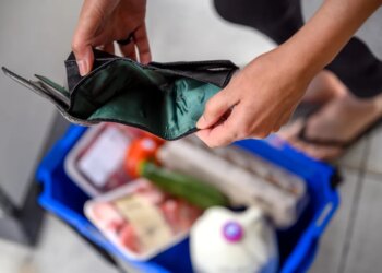 A woman holding an empty wallet over a grocery shopping cart, symbolizing financial hardship due to high food prices.