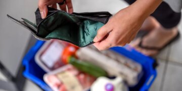 A woman holding an empty wallet over a grocery shopping cart, symbolizing financial hardship due to high food prices.