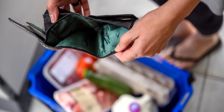 A woman holding an empty wallet over a grocery shopping cart, symbolizing financial hardship due to high food prices.