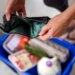 A woman holding an empty wallet over a grocery shopping cart, symbolizing financial hardship due to high food prices.