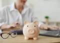A piggy bank on a desk, with a financial advisor calculating a budget in the background.