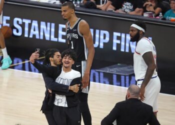 A fan taking a selfie video on the court during the 2026 NBA Finals Game 1 between the New York Knicks and San Antonio Spurs, with players looking on.