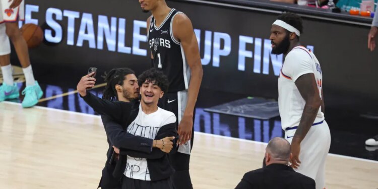 A fan taking a selfie video on the court during the 2026 NBA Finals Game 1 between the New York Knicks and San Antonio Spurs, with players looking on.
