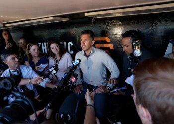 A man in a light grey shirt surrounded by reporters holding microphones.