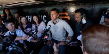 A man in a light grey shirt surrounded by reporters holding microphones.