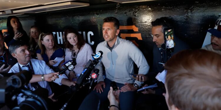 A man in a light grey shirt surrounded by reporters holding microphones.