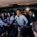 A man in a light grey shirt surrounded by reporters holding microphones.
