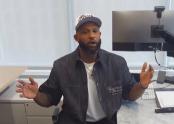 A man in a white cap, dark denim jacket, and white T-shirt sitting at a desk with a computer monitor.