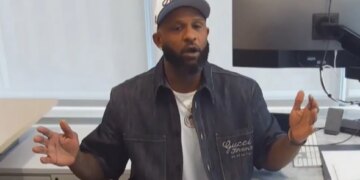 A man in a white cap, dark denim jacket, and white T-shirt sitting at a desk with a computer monitor.