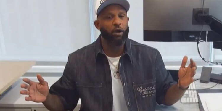 A man in a white cap, dark denim jacket, and white T-shirt sitting at a desk with a computer monitor.