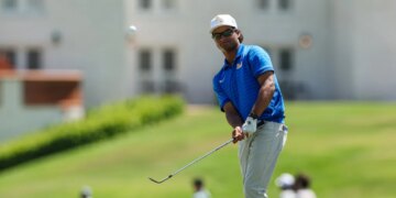 A male golfer in a blue shirt and white hat hitting a golf ball.