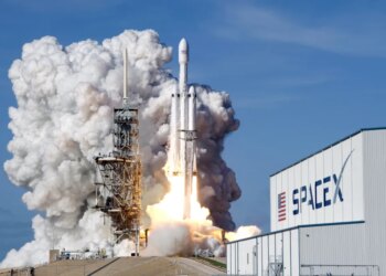 A Falcon 9 SpaceX heavy rocket lifts off from a launch pad, surrounded by a large plume of smoke and fire, with a SpaceX building visible to the right.