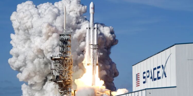 A Falcon 9 SpaceX heavy rocket lifts off from a launch pad, surrounded by a large plume of smoke and fire, with a SpaceX building visible to the right.