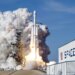 A Falcon 9 SpaceX heavy rocket lifts off from a launch pad, surrounded by a large plume of smoke and fire, with a SpaceX building visible to the right.