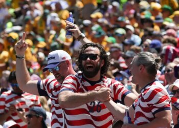 Three male US fans in striped shirts cheering at a soccer match, one holding a beer aloft.