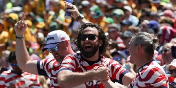 Three male US fans in striped shirts cheering at a soccer match, one holding a beer aloft.