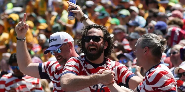 Three male US fans in striped shirts cheering at a soccer match, one holding a beer aloft.