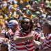 Three male US fans in striped shirts cheering at a soccer match, one holding a beer aloft.
