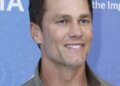 Tom Brady smiling while wearing a light brown jacket over a white shirt.
