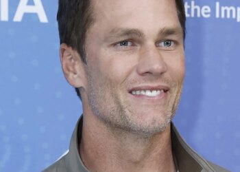 Tom Brady smiling while wearing a light brown jacket over a white shirt.