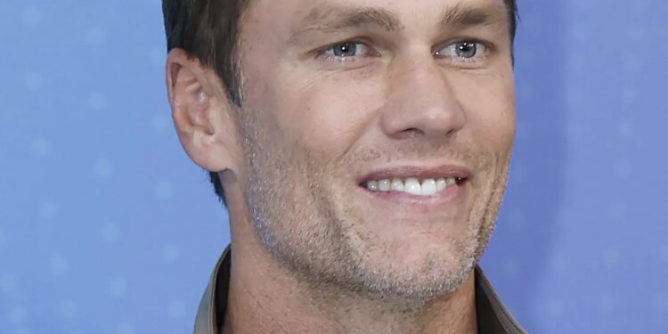 Tom Brady smiling while wearing a light brown jacket over a white shirt.