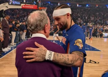 A basketball player in a blue jersey with a white headband, and an older man in a purple sweater, embracing on a basketball court.