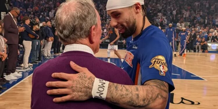 A basketball player in a blue jersey with a white headband, and an older man in a purple sweater, embracing on a basketball court.