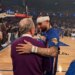 A basketball player in a blue jersey with a white headband, and an older man in a purple sweater, embracing on a basketball court.