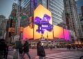 Advertising for Roku in Times Square, New York, with a prominent digital billboard featuring the Roku logo and a robot mascot against a vibrant orange and purple background, surrounded by other digital signs and buildings, and with pedestrians crossing a street in the foreground.