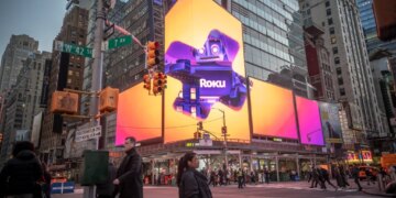 Advertising for Roku in Times Square, New York, with a prominent digital billboard featuring the Roku logo and a robot mascot against a vibrant orange and purple background, surrounded by other digital signs and buildings, and with pedestrians crossing a street in the foreground.