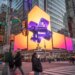 Advertising for Roku in Times Square, New York, with a prominent digital billboard featuring the Roku logo and a robot mascot against a vibrant orange and purple background, surrounded by other digital signs and buildings, and with pedestrians crossing a street in the foreground.