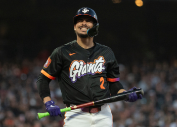 A baseball player for the San Francisco Giants, wearing a black jersey with orange and purple lettering, holds a bat and smiles.