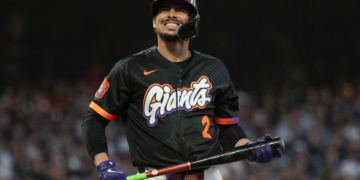A baseball player for the San Francisco Giants, wearing a black jersey with orange and purple lettering, holds a bat and smiles.