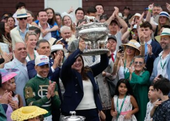 Trainer Cherie DeVaux holds the trophy aloft, flanked by jockey Jose Ortiz and cheered on by a crowd.