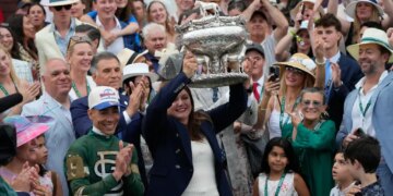 Trainer Cherie DeVaux holds the trophy aloft, flanked by jockey Jose Ortiz and cheered on by a crowd.