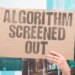A hand holding up a cardboard sign that says "ALGORITHM SCREENED OUT" in capital letters.