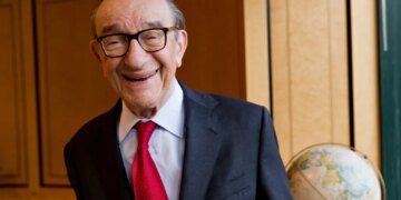 Economist Alan Greenspan smiles, wearing glasses, a suit jacket, and a red tie.