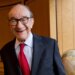 Economist Alan Greenspan smiles, wearing glasses, a suit jacket, and a red tie.
