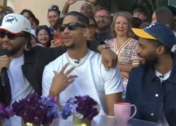 Three men are seated at a table with an audience behind them, with purple flowers and pink mugs on the table.