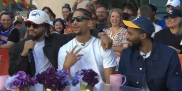 Three men are seated at a table with an audience behind them, with purple flowers and pink mugs on the table.