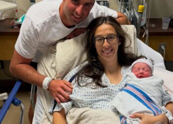 A man stands behind a woman in a hospital bed holding a swaddled newborn, all three smiling.
