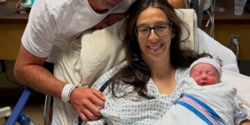 A man stands behind a woman in a hospital bed holding a swaddled newborn, all three smiling.
