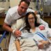 A man stands behind a woman in a hospital bed holding a swaddled newborn, all three smiling.
