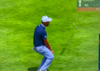 A male golfer in a white cap and blue shirt bends over, watching his golf club fly in the air after a shot, with a scoreboard visible on the right.