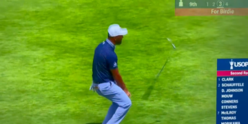 A male golfer in a white cap and blue shirt bends over, watching his golf club fly in the air after a shot, with a scoreboard visible on the right.