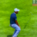 A male golfer in a white cap and blue shirt bends over, watching his golf club fly in the air after a shot, with a scoreboard visible on the right.