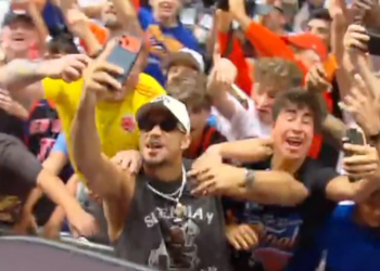 A man in sunglasses taking a selfie with a crowd of cheering people behind him.
