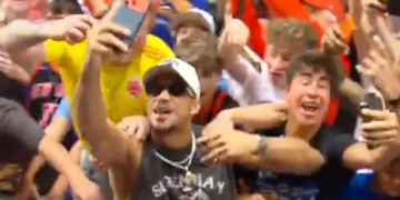 A man in sunglasses taking a selfie with a crowd of cheering people behind him.