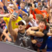 A man in sunglasses taking a selfie with a crowd of cheering people behind him.